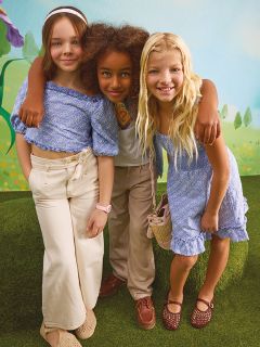 Trois jeunes filles se tiennent avec assurance au milieu des feuilles d'automne, vêtues de tenues brillantes et métalliques. Deux filles portent des vestes argentées, et la fille du milieu porte un haut et une jupe ornés de paillettes. Un fond bleu-vert contraste avec le feuillage d'automne orange.