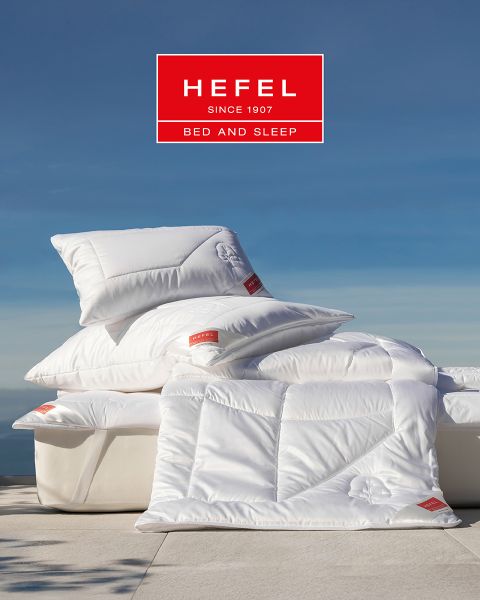 Une femme dort paisiblement sur le côté dans un lit moderne avec un oreiller beige et une couverture blanche. En arrière-plan, on aperçoit un vase avec de l'herbe de la pampa et une vue sur la montagne au coucher du soleil. En haut, on peut voir le logo de « Hefel Bed and Sleep ».