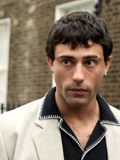 Une personne en costume marron et avec un chapeau assorti est assise sur un banc noir devant un fond rouge et blanc et regarde sur le côté avec une mine pensive.