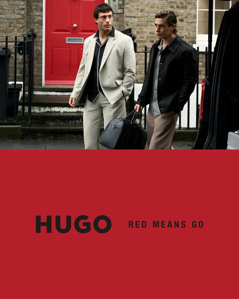 Deux mannequins en costumes noirs posent avec assurance devant un fond clair et simple. Le logo HUGO apparaît en gras et en lettres blanches dans le coin inférieur gauche.