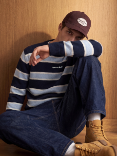 Un jeune homme en veste en jean et jean est agenouillé et regarde sur le côté. Il porte des baskets blanches et a un tatouage visible sur son avant-bras. L'arrière-plan est uni, gris clair.