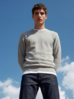 Un jeune homme portant un t-shirt noir avec l'inscription "Original Studio", un collier argenté, un bracelet argenté et une casquette de baseball rayée crème et noire se tient devant un fond gris clair uni.