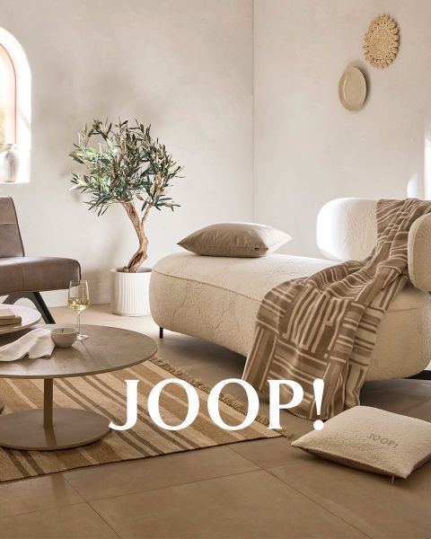 Des coussins aux couleurs neutres avec des motifs et une couverture assortie sont disposés sur un banc en béton minimaliste. À côté se trouvent un grand vase et une plante séchée décorative. L'inscription « JOOP! » figure sur la couverture. Une douce lumière du soleil projette des ombres sur la scène.