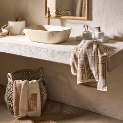 Salle de bain minimaliste avec miroir rond, robinet mural et lavabo rectangulaire. Des étagères accueillent des objets de décoration et des serviettes dans des tons terre. Une lumière douce projette des ombres légères sur le mur texturé, créant une atmosphère paisible.