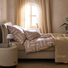Une chambre confortable avec un lit beige et du linge de lit à motifs. Un miroir rond est accroché au mur. Une lumière douce illumine une partie du lit. Une petite pile de livres et des objets de décoration sont posés à proximité sur le sol.