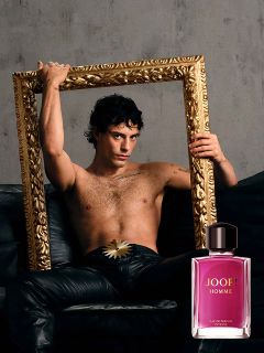 Une bouteille de JOOP! Homme Le Parfum est tenue devant un arrière-plan flou avec une fleur d'iris violette. Le logo doré de "JOOP!" est clairement visible en haut. L'image transmet élégance et luxe.