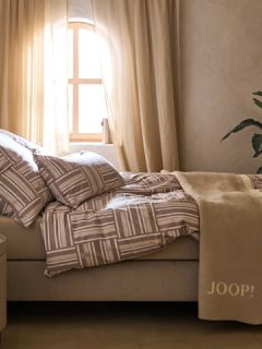 Un espace minimaliste présente un banc en pierre beige orné de deux coussins texturés bruns et beiges, d'un jeté à motifs et d'herbes séchées. Un panier tressé, un vase en céramique rustique et un grand pichet décorent le sol sous une douce lumière naturelle.