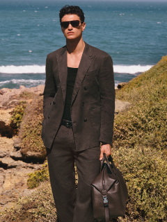 Une personne en costume noir formel se tient devant une balustrade en pierre avec des arbres en arrière-plan. Le ciel est partiellement nuageux. Sa chemise est partiellement déboutonnée et elle a une expression confiante.