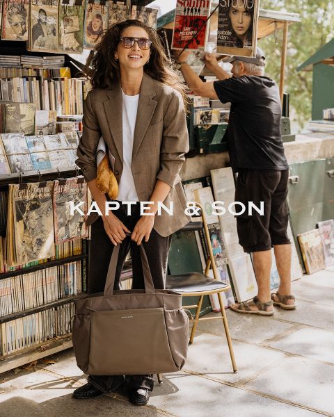 Une femme et un homme se sourient à l'extérieur. La femme porte un bonnet vert et un manteau beige et tient un sac noir à la main. L'homme porte une veste marron et un sac couleur crème. Au milieu, il y a écrit « Kapten & Son ».