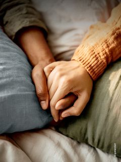 Deux personnes allongées sur un lit, se tenant la main, l'une portant une manche orange et l'autre une manche verte, pour exprimer le réconfort et la proximité.