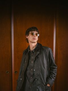 Une personne avec des lunettes de soleil et une veste sombre se tient devant des portes en bois, regarde légèrement sur le côté et a les mains dans les poches.