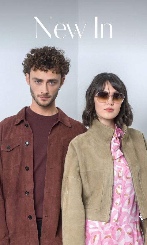 Un homme portant une veste marron et une femme portant des lunettes de soleil et une veste marron clair sur une robe à motifs roses se tiennent côte à côte devant un fond clair et uni.