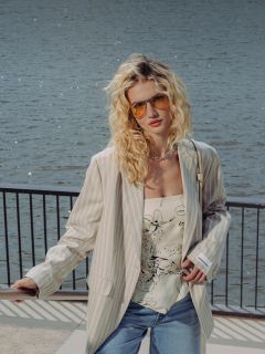 Une femme aux cheveux blonds bouclés porte des lunettes de soleil, un blazer à rayures, un haut à fleurs, un short en jean et des chaussures blanches, alors qu'elle se tient sur une promenade au bord de l'eau avec une balustrade.