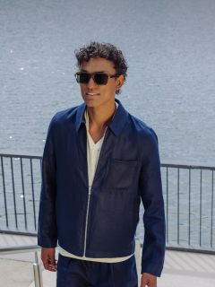 Un homme portant des lunettes de soleil, une veste bleu marine et un short assorti monte un escalier extérieur au bord de l'eau, tout en se tenant à une rampe et en souriant.