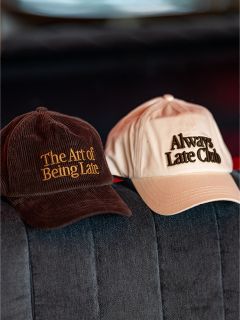 Sur un canapé gris se trouvent deux casquettes de baseball ; sur l'une, marron, est écrit « The Art of Being Late » et sur l'autre, beige, « Always Late Club ».