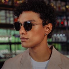 Une personne aux cheveux courts et bouclés, portant des lunettes de soleil, un blazer beige et une chemise blanche, se tient dans un intérieur avec des étagères en verre en arrière-plan.