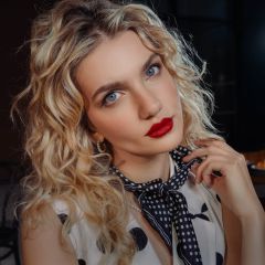 Une femme aux cheveux blonds bouclés, avec du rouge à lèvres rouge et une écharpe à pois, regarde la caméra en tenant sa main sur sa nuque.