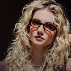 Jeune femme aux longs cheveux blonds bouclés, portant des lunettes en écaille, un collier de perles et un haut marron, regardant l'objectif.