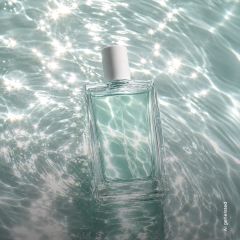 Un flacon de parfum rectangulaire en verre avec un bouchon blanc flotte sur de l'eau scintillante et ensoleillée.