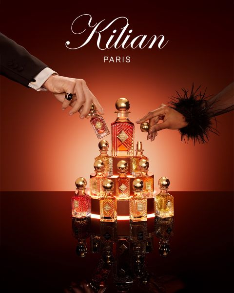 Un flacon du parfum Kilian Paris Angels' Share est posé à côté d'un verre de whisky avec des glaçons et une garniture, le tout sur une surface brune réfléchissante avec un éclairage doux. Le logo Kilian Paris est visible en haut.