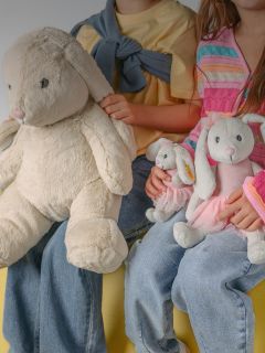 Un garçon souriant aux cheveux blonds est assis dans une maison et serre un grand ours en peluche beige dans ses bras. Il porte une chemise à manches longues noire et blanche et un pantalon clair.