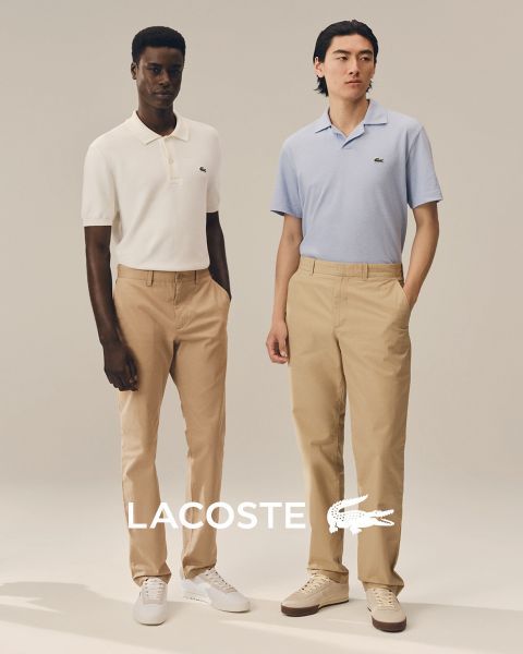 Quatre mannequins posent en extérieur, vêtus de tenues Lacoste, devant un ciel bleu. De gauche à droite : une personne en trench-coat, une personne en pull et pantalon, une personne en gilet et jupe, et une personne en survêtement. Le logo Lacoste se trouve en bas.