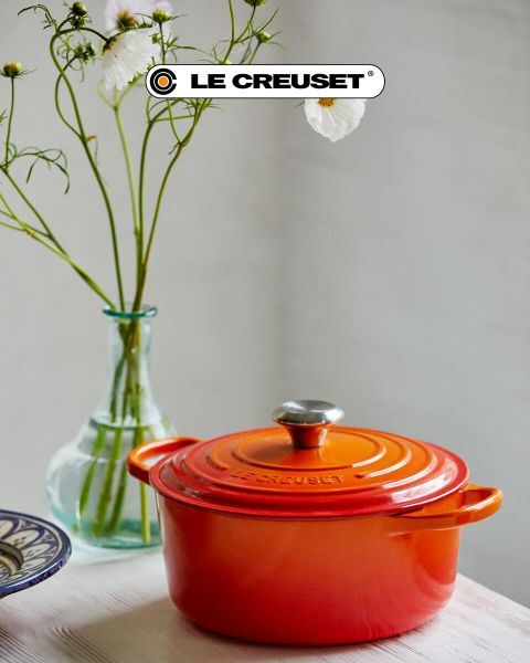 Une scène de cuisine avec une cocotte rouge, une poêle à frire antiadhésive, un ensemble de casseroles empilables en acier inoxydable, une marmite noire et un bol de tomates cerises sur un comptoir en bois clair devant un dosseret en carreaux blancs. Le logo Le Creuset est visible.