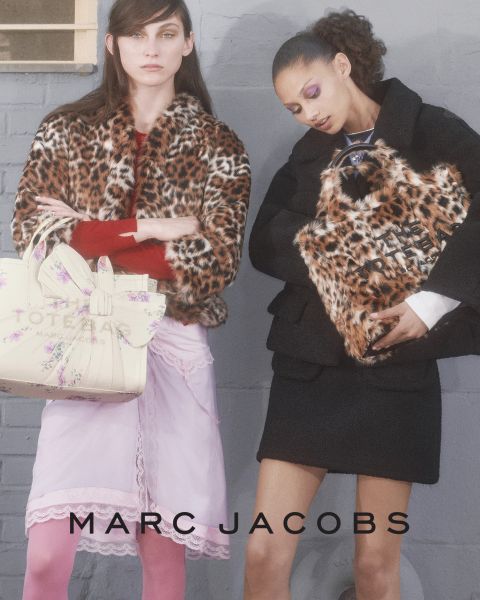 Une femme pose en extérieur avec un sac marron Marc Jacobs à la main, se penche en arrière la tête inclinée, portant une veste marron et un jean bleu. Les mots "THE TOTE BAG MARC JACOBS" et "MARC JACOBS" apparaissent sur l'image.