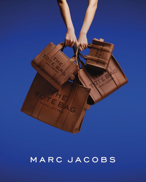Une femme pose en extérieur avec un sac marron Marc Jacobs à la main, se penche en arrière la tête inclinée, portant une veste marron et un jean bleu. Les mots "THE TOTE BAG MARC JACOBS" et "MARC JACOBS" apparaissent sur l'image.