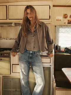Une femme aux longs cheveux blonds ondulés porte un manteau à capuche marron avec une ceinture noire, se tient devant un mur en bois rustique et regarde l'appareil photo avec assurance.