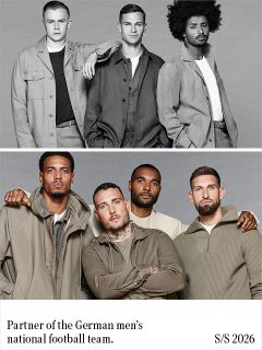 Un homme aux cheveux bouclés et foncés, portant un blazer noir sur un pull en maille côtelée verte, se tient les bras croisés devant un fond gris uni et regarde droit devant lui avec une expression neutre.