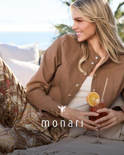 Une femme souriante portant un manteau beige, une chemise rayée, un jean bleu et une écharpe claire se tient à l'extérieur devant un grand bâtiment. Le logo Monari est visible en bas de l'image.