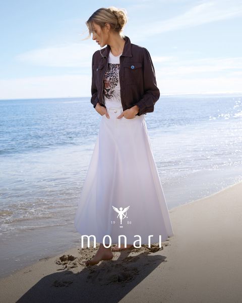 Une femme souriante portant un manteau beige, une chemise rayée, un jean bleu et une écharpe claire se tient à l'extérieur devant un grand bâtiment. Le logo Monari est visible en bas de l'image.