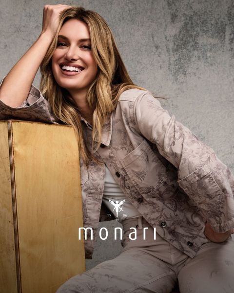 Une femme souriante portant un manteau beige, une chemise rayée, un jean bleu et une écharpe claire se tient à l'extérieur devant un grand bâtiment. Le logo Monari est visible en bas de l'image.