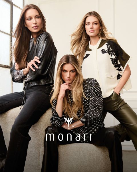 Une femme souriante portant un manteau beige, une chemise rayée, un jean bleu et une écharpe claire se tient à l'extérieur devant un grand bâtiment. Le logo Monari est visible en bas de l'image.