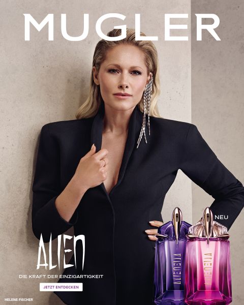 Une femme à la peau foncée et éclatante et aux cheveux noirs lisses pose devant un fond violet. Devant elle se trouvent deux flacons de parfum Alien de Mugler. Le texte indique « MUGLER » et « ALIEN EXTRA INTENSE LES FRAGRANCES RECHARGEABLES ».