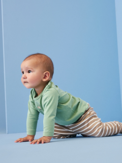 Un bébé souriant est allongé sur une couverture blanche, portant un pull en tricot bleu, un pantalon en velours côtelé marron avec des poches et des chaussettes grises. Le bébé a l'air heureux et détendu, avec les bras légèrement pliés au niveau des coudes.