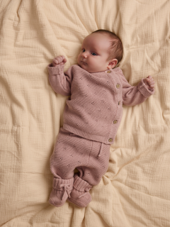Un bébé est allongé sur une couverture blanche et porte un pull en tricot rose clair avec un col Claudine et un pantalon assorti, ainsi que des chaussettes couleur crème.