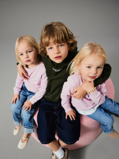 Deux jeunes enfants se tiennent par la main devant un fond marron uni. Un enfant porte une veste rose et une jupe, l'autre un jean bleu et une chemise bleue avec l'inscription "L'AMOUR EST ENFANT". Les deux regardent vers le haut avec curiosité.