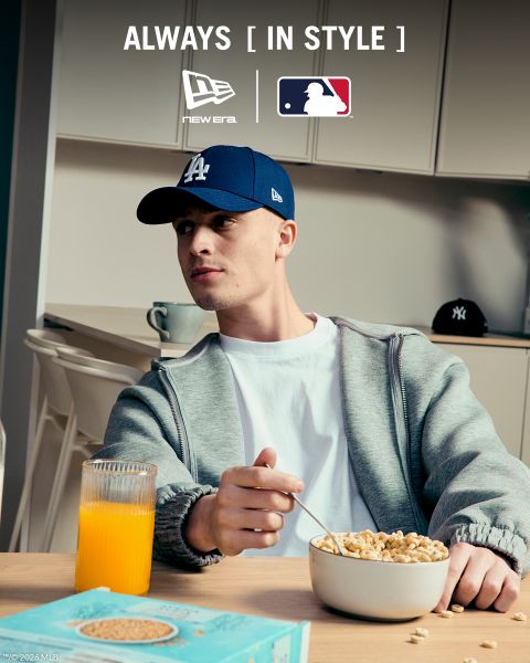 Un homme portant un survêtement gris, un t-shirt blanc et une casquette bleu marine des New York Yankees est assis sur un banc en bois et écoute de la musique avec des écouteurs blancs. Le texte fait la promotion de la casquette 9FORTY M-Crown de New Era avec la marque MLB.