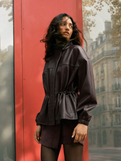 Une femme portant un costume marron oversize avec une ceinture noire et des bijoux en or traverse une rue en ville avec assurance, portant un grand sac à main noir. En arrière-plan, on aperçoit des clients de café assis.