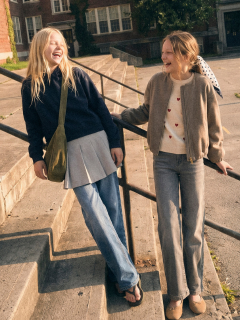 Deux jeunes filles sourient et dansent ensemble dans une maison. L'une porte un t-shirt noir scintillant et une jupe, l'autre un cardigan foncé sur une robe. On aperçoit des ballons métalliques et des étagères rouges en arrière-plan.