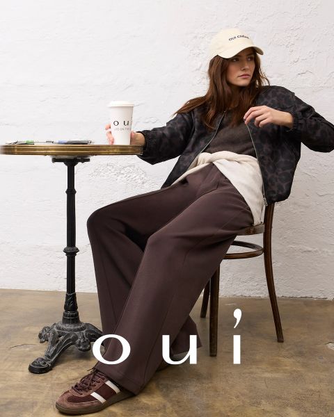Une femme portant un pull rouge avec un cœur blanc, un jean bleu et un bonnet blanc pose devant un mur clair. En bas de l'image, le mot « oui » est écrit en lettres blanches.