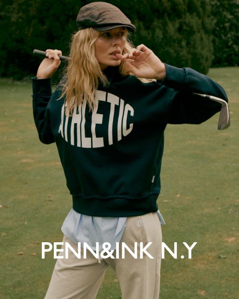 Une femme aux longs cheveux ondulés porte un tailleur foncé et des sandales à talons hauts, et se tient avec assurance dans un studio avec un fond uni. Le texte en bas indique "PENN&INK N.Y."