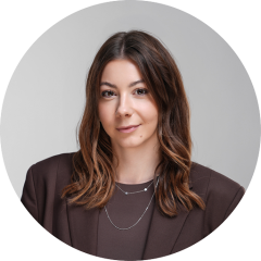 Femme aux longs cheveux bruns, portant un blazer marron et des colliers superposés, et souriant doucement devant un fond gris neutre.