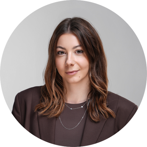 Femme aux longs cheveux bruns, portant un blazer marron et des colliers superposés, et souriant doucement devant un fond gris neutre.