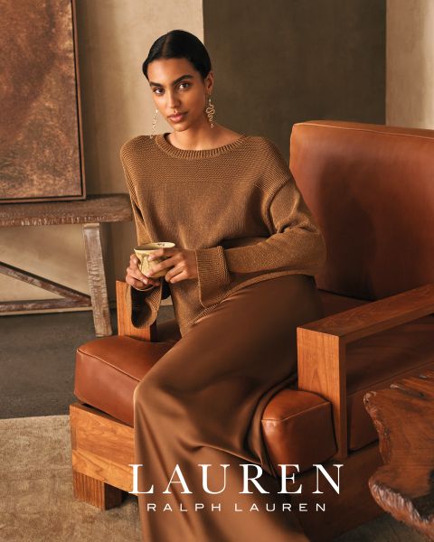 Une femme aux longs cheveux ondulés est appuyée sur un canapé blanc, portant un haut rouge vif et un pantalon assorti. Une lumière chaude du soleil projette des ombres sur la scène. En bas, on peut lire "LAUREN RALPH LAUREN".
