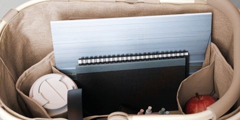 Une personne aux longs cheveux roux, portant une chemise bleu clair et un pantalon blanc, se détend dans l'herbe et tient un sac à motifs à la main. Elle se trouve à l'extérieur, dans un champ ensoleillé, avec vue sur les collines au loin. Le mot "Reisenthel" est visible en rouge.