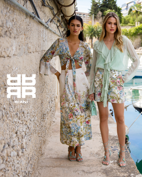 Deux femmes posent avec style dans un théâtre aux sièges rouges. L'une est assise sur le dossier d'un siège et porte une robe à motifs, un manteau noir et des bottes. L'autre se tient à côté d'elle en pull bleu marine et jupe noire. Au-dessus d'elles apparaît le logo "RIANI".