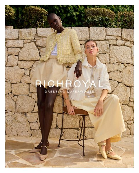 Une femme en chemise bleu clair et pantalon gris tient une boule à neige devant un œil. Elle pose dans une pièce aux murs en bois avec une affiche de ski et porte de grandes boucles d'oreilles. Le texte est "RICHROYAL DRESSING SUPERWOMEN".